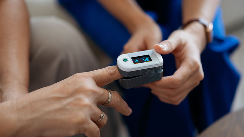 Pessoa a medir a saturação de oxigénio no dedo com um oxímetro portátil, enquanto outra pessoa observa o visor do dispositivo.